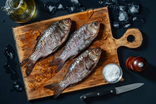 Fresh raw fish crucian carp with ingredients on wooden cutting board, close up. Top view of three raw silver fish on a wooden board with ice cubes, olive oil, salt, pepper and knife