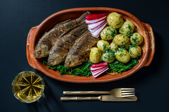 Fried river fish with boiled baby potatoes and dill in clay dish, close up. Top view of crispy fried fish served with boiled young potatoes, greens, radish and drink on black background