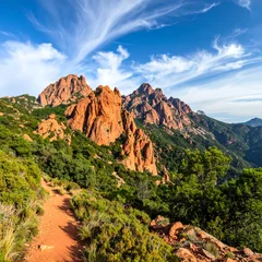 A serene mountain trail surrounded by lush greenery and vibrant red rock formations under a bright blue sky with wispy white clouds © Karang