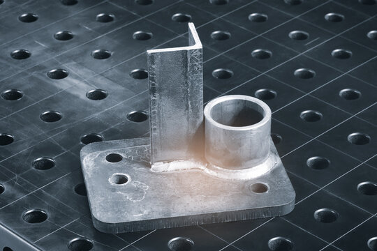 Metalwork components rest on black grid surface. Cylindrical tube and L-shaped bracket show machining marks. Base plate has rusted edges and mounting holes