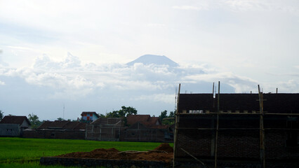 Obraz premium Volcanic mountain peak emerging through clouds above rural village with unfinished house scaffolding and lush green rice fields in foreground.