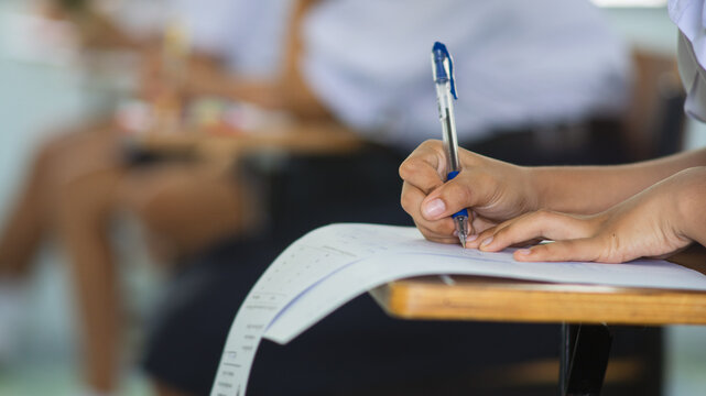 Close-up of a student's hand diligently writing on an exam paper during a school test. The student is using a professional pen , sitting at a wooden desk in a brightly lit classroom. 