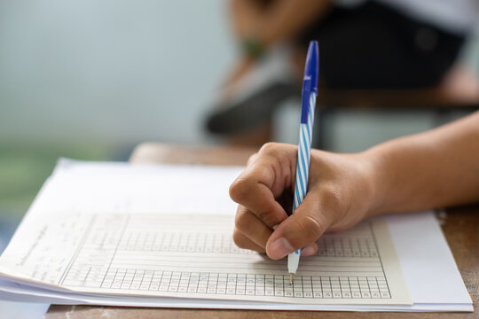 Close-up of a student's hand diligently writing on an exam paper during a school test. The student is using a professional pen , sitting at a wooden desk in a brightly lit classroom. 