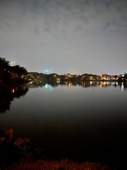 Fototapeta premium Night landscape of a calm lake reflecting a house and white clouds on the water surface. Mirror-like reflection creates a peaceful scenic image with symmetry, tranquility, and natural beauty.