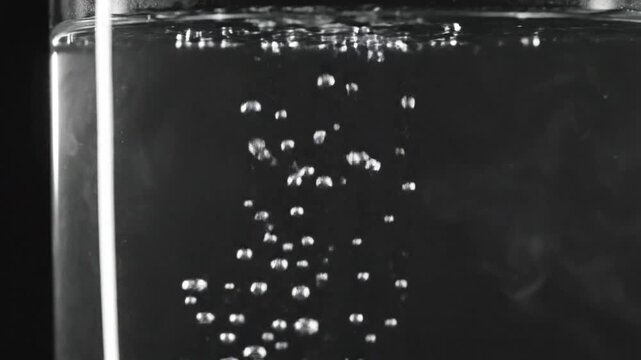 Boiling water in a glass, vapor rising, steam bubbles in transparent liquid on black background