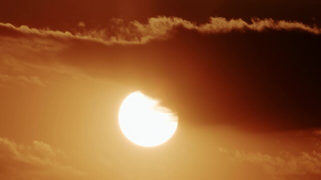 Close up of a bright sun partially covered by thick moving clouds, glowing in intense golden orange sunset tones