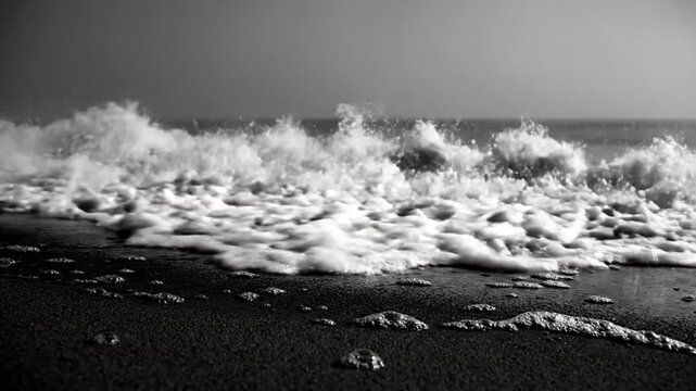Black and white close up of ocean waves crashing onto sandy beach creating sea foam