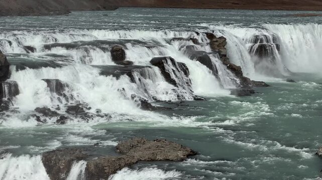 Gullfoss Waterfall flows into river valley, surrounded by cliffs and snow patches, showcasing Iceland's breathtaking natural beauty and rugged terrain