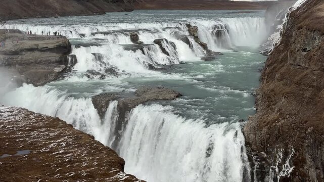 Gullfoss Waterfall flows into river valley, surrounded by cliffs and snow patches, showcasing Iceland's breathtaking natural beauty and rugged terrain