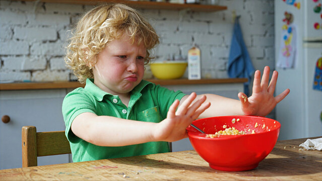 Young Blonde Boy with Curly Hair Refusing Food, Pushing Away Red Bowl with Disgusted Facial Expression at Kitchen Table, Picky Eater and Toddler Mealtime Tantrum Concept