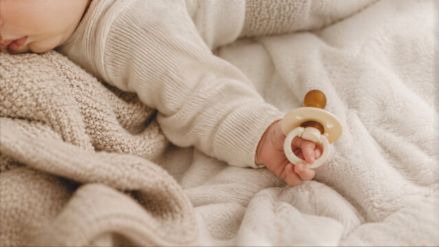Close-up of a peaceful newborn baby sleeping on a soft beige blanket holding a natural rubber pacifier, detailed shot of infant's tiny hand and cozy knit sweater.