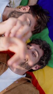 Intimate moment of gay couple on Pride flag, eyes locked with aﬀection, sharing a
smile that speaks volumes of their bond. Two men in love, exchange joyful glances,
tenderly touching each other hands