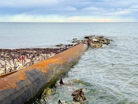 An old rusted metal pipeline extending from the sandy shore into the choppy waters of the Baltic Sea in Latvia