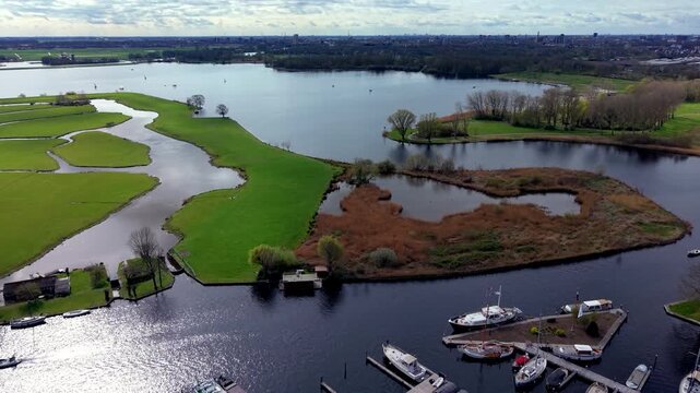 Aerial drone view over a marina harbor with boats at the docks, adjacent to a lush green polder landscape, winding waterways, and islands leading to a large lake.