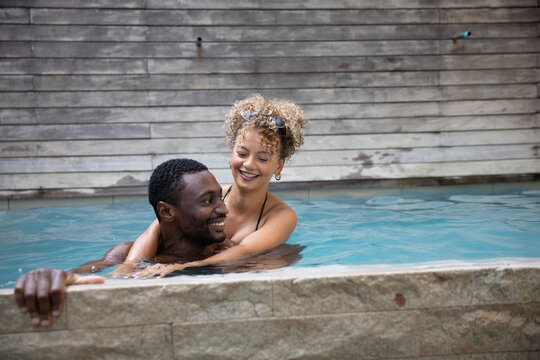 Diverse couple wearing bikini hugging while swimming at outdoor pool near stone coping, wooden wall