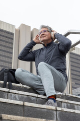 Senior man sitting on stone steps in city plaza adjusting headset in athletic wear with backpack © wavebreak3