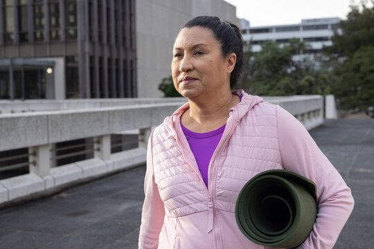 Woman holding rolled green yoga mat under left arm, walking on raised walkway wearing pink hoodie