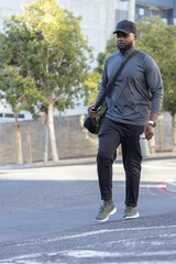 Obraz premium Walking on pavement, African man wearing cap and pullover, carrying crossbody bag and water bottle