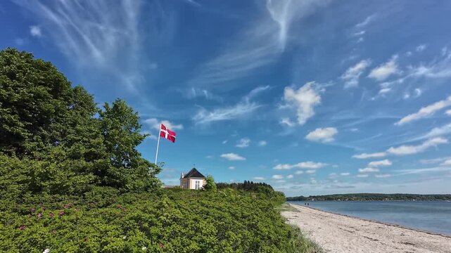 Scenic wide video of the historic yellow tea house pavilion at Valdemars Slot on Tasinge with the national Dannebrog flag waving in the wind by a beach under a beautiful sky