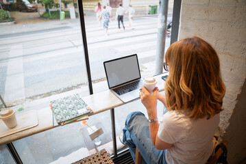 Mature woman working remotely from coffee shop with laptop and takeaway coffee
