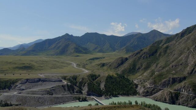 Drone footage of the Chuysky Trakt highway winding through a wide mountain valley with rocky slopes and a turquoise river in Altai Republic, Russia