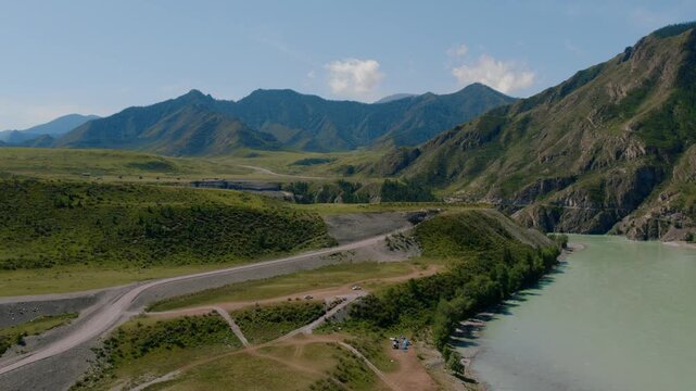 Drone footage of Chuysky Trakt highway winding through green hills beside the turquoise Katun River in the Altai Republic, Russia, on a clear summer day