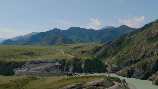 Drone footage of the Chuysky Trakt road curving through a wide green valley with rugged mountains and a turquoise river in Altai Republic, Russia