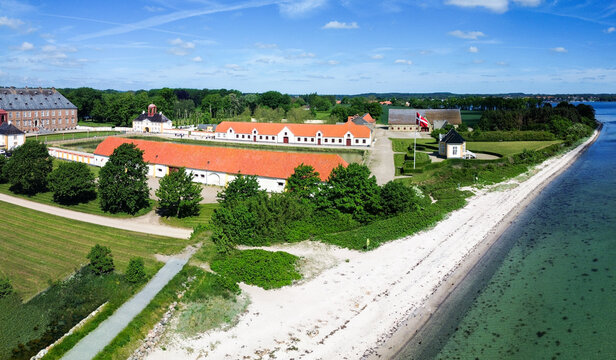 Valdemars Slot. The national flag of Denmark, Dannebrog, waves proudly on a flagpole near a white sand beach and calm coastal waters on a sunny summer day