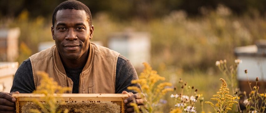 Black beekeeper holding honeycomb frame in apiary during golden hour, smiling African American apiarist working at honey bee farm with wooden hives, sustainable organic agriculture and apiculture.