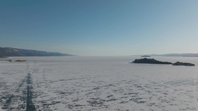 Wide aerial view of frozen Lake Baikal with textured ice, cracks, and tracks, framed by distant mountains under clear blue sky, Russia, Siberia in winter