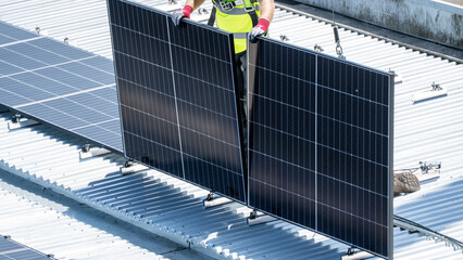 Worker in yellow vest holding two large solar panel upright on rooftop © Preethiloki
