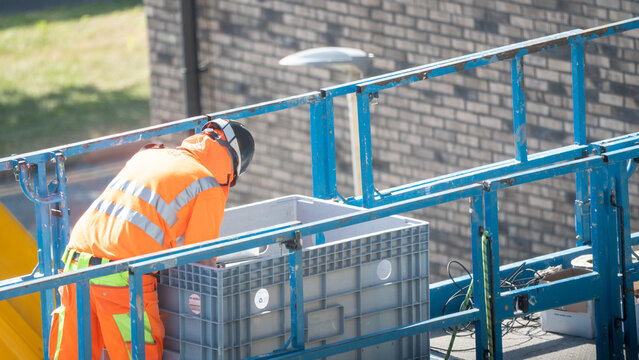 Worker in orange safety vest bending over grey crate on blue scaffold platform