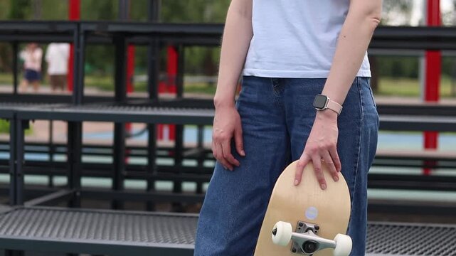 Girl in blue jeans and white t-shirt holding skateboard, standing against city tribunes background. Young girl with skateboard on sports ground. Modern street style and active leisure