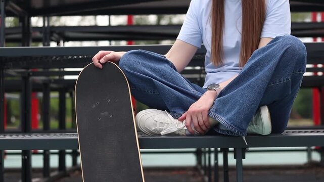 Young girl in baggy jeans sitting on metal bleachers next to skateboard. Close-up of teenage girl sitting in lotus position at stadium with skateboard. Skater resting at city sports ground