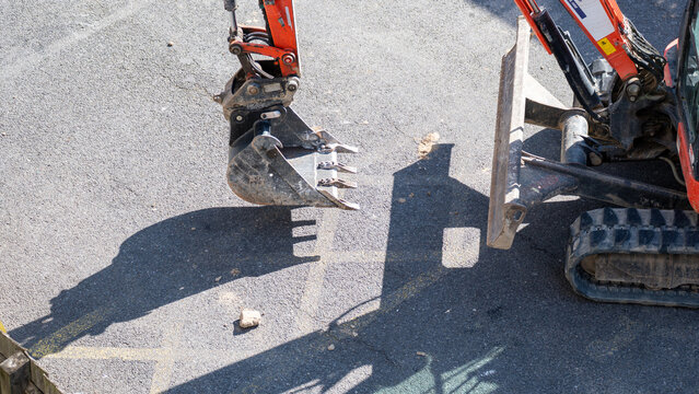 Mini excavator with metal bucket parked on gravel casting shadow on paved ground
