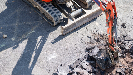 Mini excavator digging into broken asphalt pavement with bucket on road surface © Preethiloki
