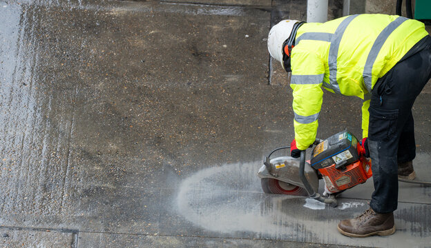 Construction worker in yellow vest cutting wet concrete road with power circular saw