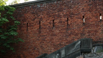 Monumentalny mur obronny z ciemnoczerwonej cegły z widocznymi otworami strzelniczymi, stanowiący fragment zabytkowej fortecy obronnej w otoczeniu zielonych liści drzew w letni dzień © tomo81