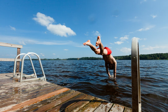 Man dives off pier with arms spread entering lake water on sunny summer day. Diver in red trunks descends with confident form and spread arms outdoors. Athletic guy creates splash with bold plunge