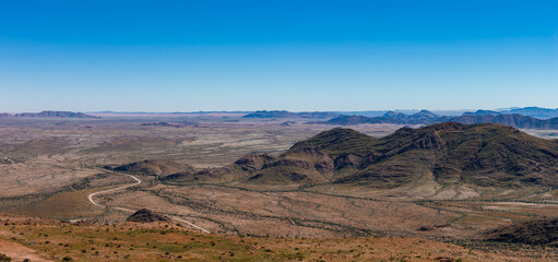 Obraz premium Aerial view of a semi arid Namibia landscape with low dark hills and mesas, a winding dirt road, and vast gravel plain under clear daylight, evoking solitude and scale.