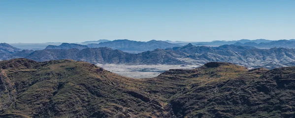Aerial view shows slate blue ridges rolling to a pale sandy basin in Namibia, Namib Desert area. Bright daytime light, long horizons, layered ranges, and a dry valley floor. © Aerial Film Studio