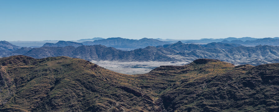 Aerial view shows slate blue ridges rolling to a pale sandy basin in Namibia, Namib Desert area. Bright daytime light, long horizons, layered ranges, and a dry valley floor.