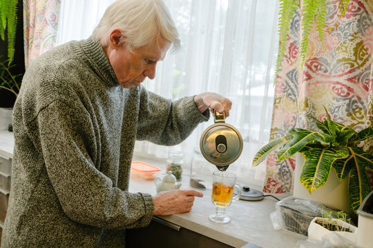 A senior citizen pours tea by himself, emphasizing theme of solitude in old age. Quiet kitchen scene speaks to experience of living alone. Grandpa in contemplative moment.