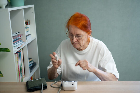 A mature woman arranges a blood pressure monitor and its components on a table at home, getting everything ready for use. Theme of health preparedness.