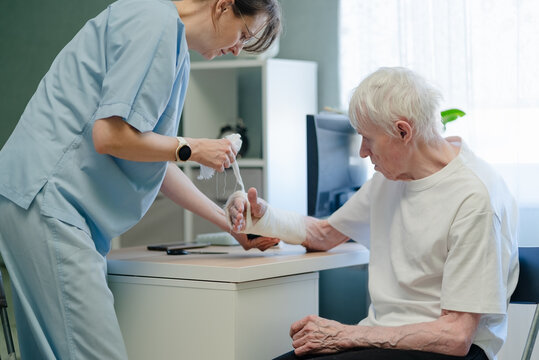 Nurse assisting elderly man with wrist bandage in clinic. Healthcare professional providing supportive medical care and gentle attention to senior patient with injured hand indoors