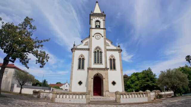 A symmetrical wide angle shot of the white facade of the historic parish church in Santa Maria de Lamas under a bright blue sky with wispy clouds. The bright daylight accentuates the textured stone de