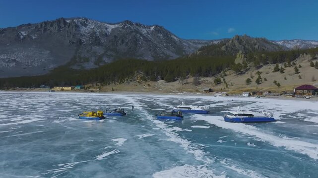 Drone footage of boats and hovercraft on cracked blue ice near the sandy shore of Peschanaya Bay, Lake Baikal, Irkutsk Oblast, Russia on a sunny winter day