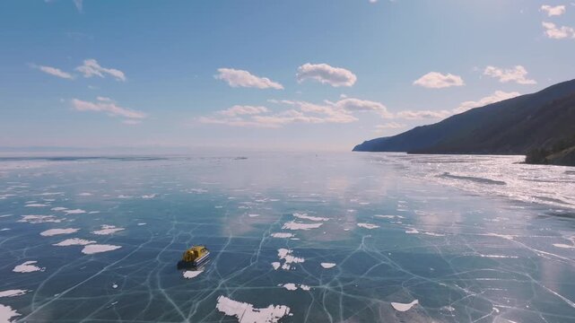 Aerial footage of a hovercraft traveling across the cracked blue ice of Lake Baikal in winter, with snowy shoreline and mountains under a clear sky, Russia