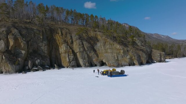 Drone footage of frozen Lake Baikal with rocky shoreline, taiga forest and mountain ridge under blue sky, winter landscape in Irkutsk Oblast, Russia