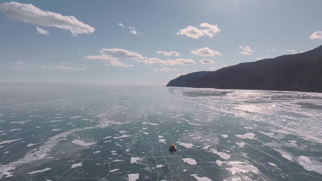 Aerial winter footage of a hovercraft traveling across frozen Lake Baikal with clear ice, cracks and snow patches, mountains on the shore, Irkutsk Oblast, Russia